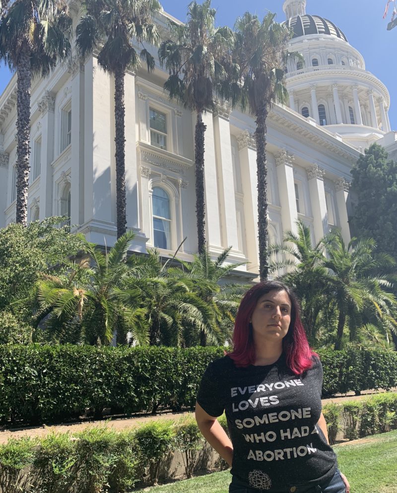 NNAF individual member Shireen wears a black t-shirt that reads, "Everyone Loves Someone Who Had an Abortion," stands in front of a bright white building with classical columns and palm trees. 