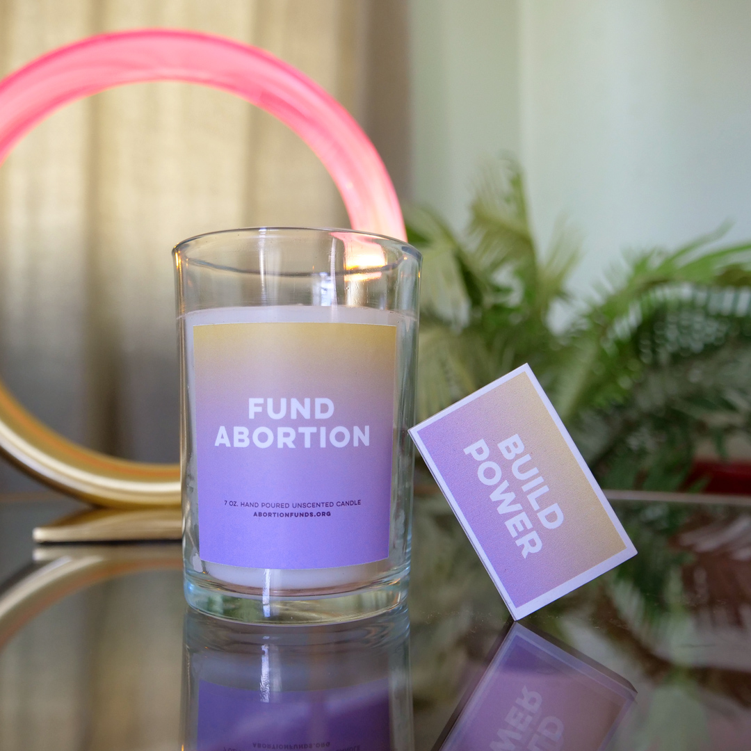 A candle in a small, clear, thick glass jar, with a label that fades from pale yellow to lavender, featuring the phrase FUND ABORTION in a clean modern sans serif font. A box of matches leans against the candle at an angle, and reads BUILD POWER, in a matching font on a matching gradient background. In the background, there's a circular lamp in brass with glowing pink lucite, and a fern.